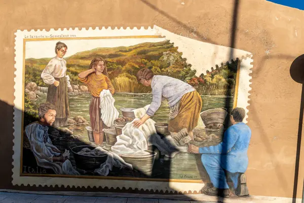 Semestene, Sardinya - İtalya - 01-12-2025: Bir duvar resmi, metal havzalarda çamaşır yıkayan nehir kenarındaki geleneksel çamaşırcı kadınları tasvir ediyor. Tablolarda bej bir duvara resim çizen bir adam var..