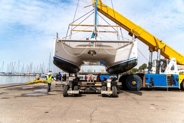 Cagliari - Sardinien - Italien - 03-14-2025: Büyük sarı bir vinç, arka planda operasyon ve marinayı yöneten işçilerin bulunduğu bir tersanede bir katamaranı sudan çıkardı.