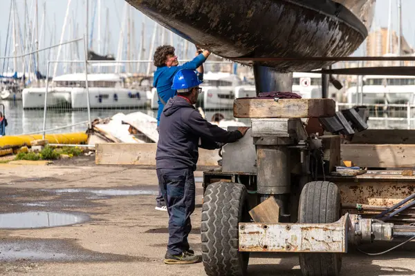 Cagliari - Sardinien - Italien - 03-14-2025: İki işçi rıhtımdaki bir marina dolusu teknenin yakınındaki bir karavanda büyüyen katamaranın bakımını ve bakımını yapıyor.