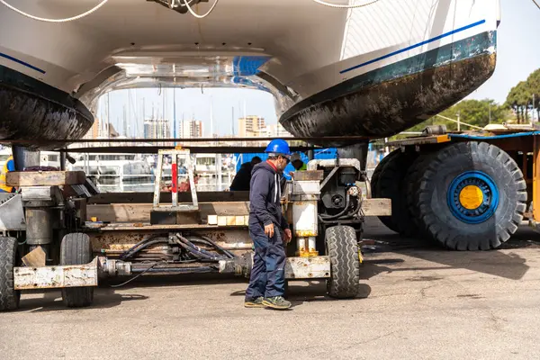 Cagliari - Sardinien - Italien - 03-14-2025: Mavi kasklı bir işçi, gündüz koşullarında bir marinada römork ve vinç destekli bir katamaranın yanında duruyor.