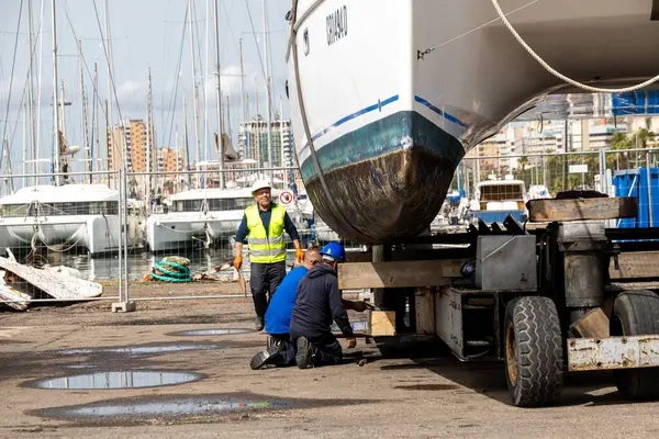 Cagliari - Sardinien - Italien - 03-14-2025: Güvenlik yelekleri içindeki üç işçi, arka planda tekneleri olan bir marinada yükseltilmiş bir katamaranın altındaki destek yapılarını düzeltiyor.
