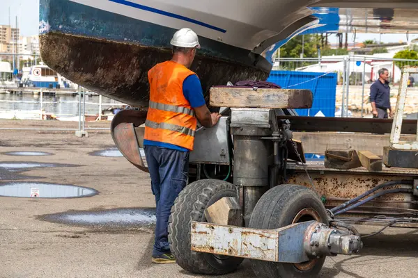 Cagliari - Sardinien - Italien - 03-14-2025: Yansıtıcı yelek giyen işçi, paslı tekerlekleri ve yapısal destekleri olan katamaran altındaki tekne römorkunun yanında kontrolleri yürütüyor.