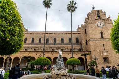 Monreale - Sizilien - Italien - 03-18-2025: Ziyaretçiler, saat kulesi ve kemerlerin yer aldığı tarihi bir katedralin önündeki budanmış ağaçlarla dolu bir avluda toplandılar..