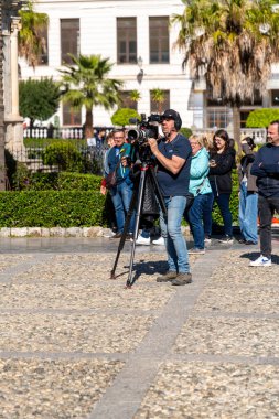 Palermo - Sizilien - Italien - 03-18-2025: Kameraman güneşli bir günde çekimler sırasında arka planda mürettebat ve palmiye ağaçlarıyla birlikte tripoda büyük bir video kamera kullanıyor.