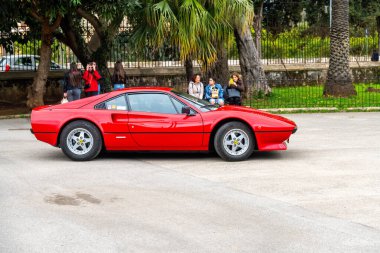 Palermo - Sizilien - Italien - 03-19-2025: Palermo caddesinde park etmiş kırmızı klasik bir spor araba, insanlar yakınlarda toplanmış, arka planda palmiye ağaçları ve demir çitlerle çerçevelenmiş..