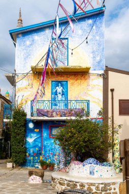 Parrini - Sizilien - Italien - 03-20-2025: Renkli duvar resimleri, mozaik dekorasyonlar, mavi balkon korkulukları ve bulutlu gökyüzü altında şenlikli flamalar bulunan canlı iki katlı bir ev.