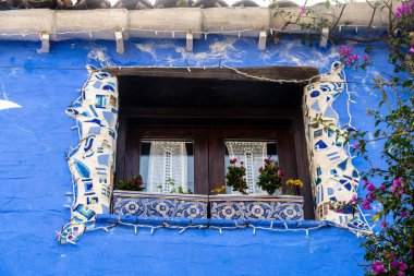 Parrini - Sizilien - Italien - 03-20-2025: Renkli mozaik fayanslarla çerçevelenmiş dekoratif ahşap pencere, canlı mavi boyalı bir duvar, çiçek saksıları ve bitkilerle çevrili.