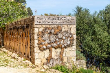 Scopello - Sizilien - Italien - 03-21-2025: Sardinya 'daki Zingaro Rezervi' nin taş giriş duvarı, metal palmiye ağaçları heykelleri ve açık mavi gökyüzü altında kırsal bir İtalyan hoş geldin tabelası.