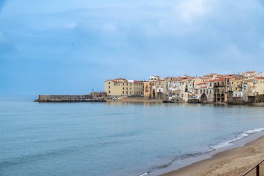 Cefalu - Sizilien - Italien - 03-24-2025: Cefalu kasabasında bulutlu bir gökyüzünün altında sakin mavi deniz ve kumlu plaj boyunca kırmızı çatıları olan kıyı binaları kümesi.