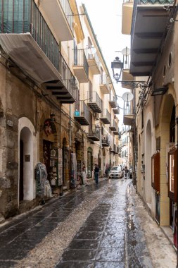 Cefalu - Sizilien - Italien - 03-24-2025: Cefalu, Sicilya 'da son yağmurdan sonra eski binalar, balkonlar, hediyelik eşya dükkanları, yayalar ve park edilmiş araçlar içeren ıslak kaldırım taşı sokak.