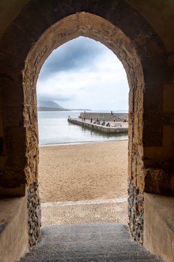 Cefalu - Sizilien - Italien - 03-24-2025: Sicilya, Cefalu 'da bulutlu bir gökyüzünün altında, kumlu bir sahilde yürüyen insanlar ve sakin bir deniz manzarası.