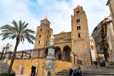 Cefalu - Sizilien - Italien - 03-24-2025: Cefalu Katedrali 'nin ön manzarasında ikiz kuleler, kemerli giriş, taş heykeller ve palmiye ağaçları ve tarihi binaların yakınında yürüyen insanlar görülüyor.