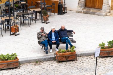 Cefalu - Sizilien - Italien - 03-24-2025: Bulutlu bir günde Sicilya 'nın Cefalu kentindeki taş döşeli Domplatz' daki boş kafe masalarının yakınındaki bir bankta üç kişi oturuyor.