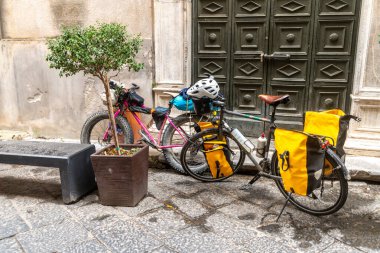 Cefalu - Sizilien - Italien - 03-24-2025: Sarı pannier taşıyan iki dolu bisiklet Cefalu, Sicilya 'da bir bank ve fabrikanın yanındaki taştan bir ara sokakta koyu yeşil bir kapıya park edildi.
