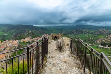 Sperlinga - Sizilien - Italien - 03-25-2025: Bulutlu bir günde Sicilya 'nın Burg Sperlinga kentindeki yeşil bir vadi ve köye bakan güçlendirilmiş bir kemer yoluna çıkan demir parmaklıklı taş geçit.