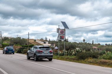 Niscemi, Sizilien - İtalya - 03-26-2025: 48 km / saat 'lik hız göstergesini geçen iki araba, ev, bitki örtüsü ve bulutlu gökyüzü ile kaplı kırsal bir yolun yanındaki güneş direğine monte edildi.