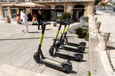 Ragusa, Sizilien - İtalya - 04-03-2025: Ragusa 'da bir restoran ve renkli vazoların yakınındaki taş bir meydanda düzgün park edilmiş sarı işaretli beş siyah elektrikli scooter.