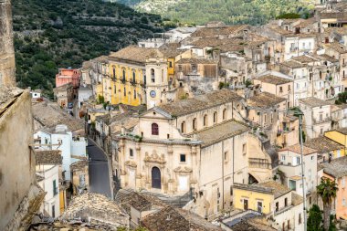 Ragusa, Sizilien - İtalya - 04-03-2025: Ragusas mimarisinin yakın çekim görüntüsü, tarihi binalar, çatılar ve bulutlu bir gökyüzünün altında bir saat kulesi.