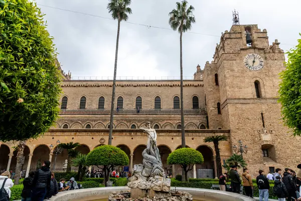 Monreale - Sizilien - Italien - 03-18-2025: Ziyaretçiler, saat kulesi ve kemerlerin yer aldığı tarihi bir katedralin önündeki budanmış ağaçlarla dolu bir avluda toplandılar..