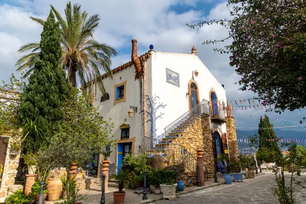 Parrini - Sizilien - Italien - 03-20-2025: Taş merdivenli, mavi seramik süslemeli Akdeniz evi, bereketli bitkiler, palmiye ağaçları ve parçalı bulutlu gökyüzü altında renkli bayraklar.