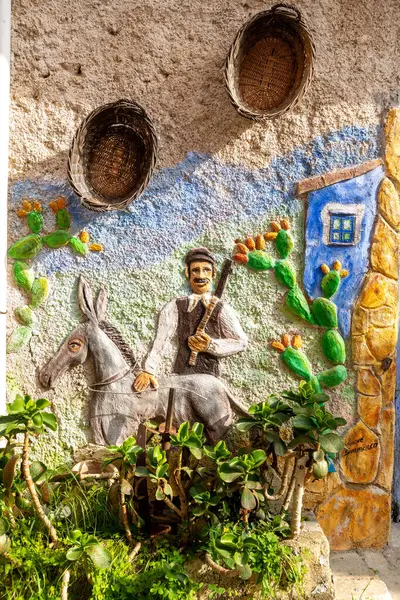 Parrini - Sizilien - Italien - 03-20-2025: Eşeğe binen, kaktüs bitkileri, dokulu duvarlar ve güneş ışığı altında asılı sepetlerle çevrili bir çiftçiyi resmeden renkli bir duvar kabartması.