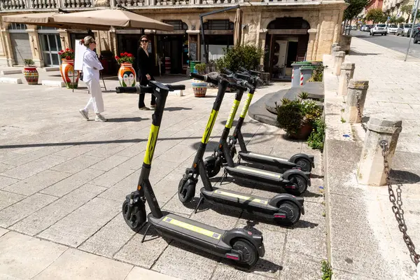 Ragusa, Sizilien - İtalya - 04-03-2025: Ragusa 'da bir restoran ve renkli vazoların yakınındaki taş bir meydanda düzgün park edilmiş sarı işaretli beş siyah elektrikli scooter.