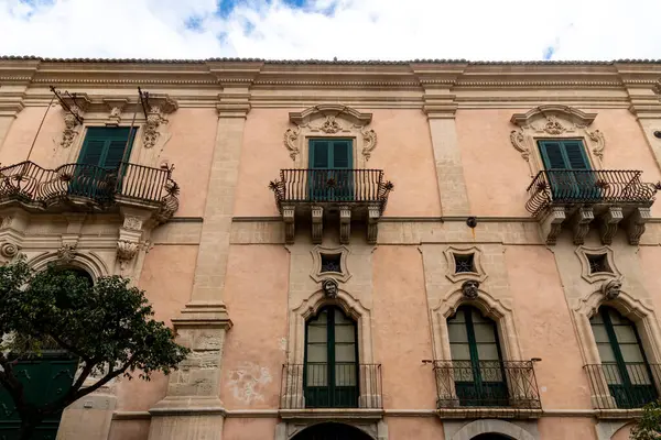 Ragusa, Sizilien - İtalya - 04-03-2025: Süslü balkonları, yeşil kepenkleri, oyulmuş taş detayları ve Sicilya 'nın tarihi merkezinde pastel duvarları olan Barok cephesi.