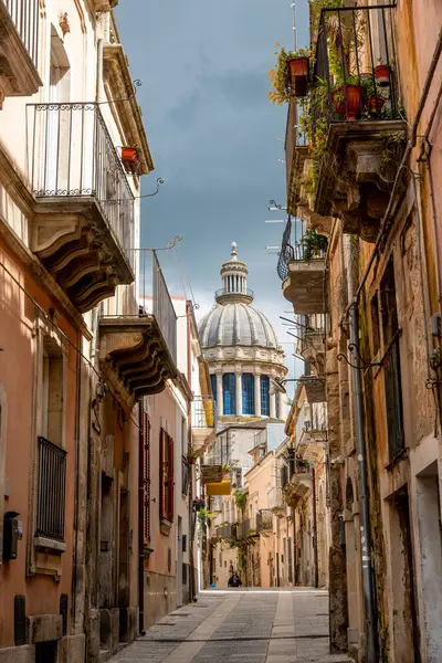 Ragusa, Sizilien - İtalya - 04-03-2025: Ragusa, Sicilya 'da bulutlu bir gökyüzünün altında mavi pencereli büyük bir katedral kubbesine uzanan eski bina ve balkonlarla dolu dar bir sokak.