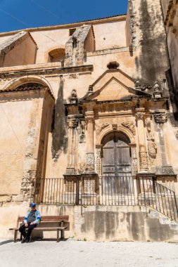 Scicli, Sizilien - İtalya - 04-05-2025: Scicli 'deki Santa Maria della Consolazione cephesinde ayrıntılı taş sütunlar, süslü kapılar ve açık mavi gökyüzünün altında bir bankta oturan bir adam görülüyor.