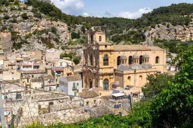 Scicli, Sizilien - İtalya - 04-05-2025: Santa Maria La Nova Kilisesi, Scicli 'deki kayalık kayalıklar ve yemyeşil gökyüzü altında parçalı bulutlu bir yay altında yer alan kiremitli çatıların üzerinde duruyor.