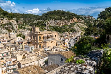 Scicli, Sizilien - İtalya - 04-05-2025: Santa Maria La Nova Kilisesi, Scicli 'deki kayalık kayalıklar ve yemyeşil gökyüzü altında parçalı bulutlu bir yay altında yer alan kiremitli çatıların üzerinde duruyor.