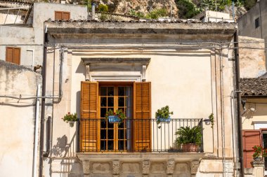 Scicli, Sizilien - İtalya - 04-05-2025: Scicli 'de demir parmaklıklar ve saksı bitkilerinin yer aldığı küçük balkon, açık ahşap panjurlar, hafif taş cephe ve açık mavi gökyüzü.