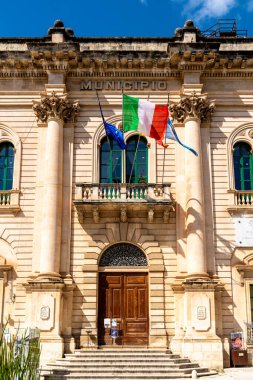 Scicli, Sizilien - İtalya - 04-05-2025: Scicli 'deki tarihi belediye binasının ön görüntüsü, taş cephe, ahşap kapılar ve açık mavi gökyüzü altında bayraklar sallıyor.