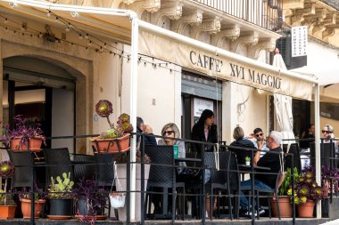 Noto, Sizilien - İtalya - 04-06-2025: Misafirler, saksı bitkileri ve sicim lambalarıyla çevrili, açık bir kafede oturmuş, sohbet ediyor ve içeceklerin keyfini çıkarıyorlar.