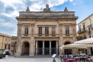Noto, Sizilien - İtalya - 04-06-2025: Süslü cepheli, heykeli ve sütunlu tarihi tiyatro Noto 'da bir meydanda, açık oturaklı ve beyaz şemsiyeli insanlar.