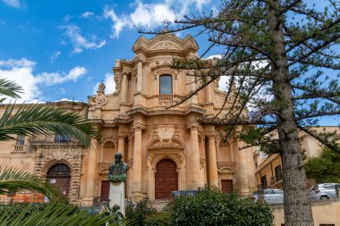 Noto, Sizilien - İtalya - 04-06-2025: Sütunları ve kavisli kapıları olan barok kilise cephesi, bir heykel, palmiye yaprakları ve mavi gökyüzünün altında bulutlu çam dalları ile çevrili.