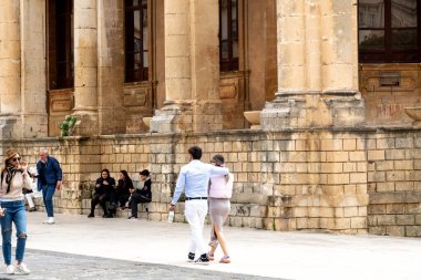 Noto, Sizilien - İtalya - 04-06-2025: Sütunları ve taş duvarları olan büyük tarihi bir binanın yanında yürüyen ve oturan insanlar, sohbete daldılar veya mola verdiler.