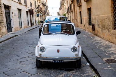 Noto, Sizilien - İtalya - 04-06-2025: Noto 'da dar bir kaldırım taşı caddesinde park etmiş klasik beyaz Fiat 500, yolun her iki tarafında da tarihi binalar bulunuyor.
