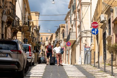 Noto, Sizilien - İtalya - 04-06-2025: Sırt çantalı ve bavullu iki turist Noto 'da dar bir sokakta yokuş yukarı yürüyor, park halindeki arabalar ve eski binalarla çevrili..