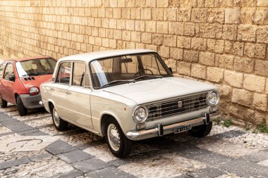 Noto, Sizilien - İtalya - 04-06-2025: Açık renkli vintage Fiat sedan taştan bir duvarın yanındaki eğimli kaldırım taşı caddesinde park edilmiş, krom detaylar ve retro tasarım sergileniyor.