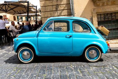 Noto, Sizilien - İtalya - 04-06-2025: Arkasında hasır sepet olan Noto 'da canlı bir açık kafenin yanındaki kaldırım taşı caddesinde park edilmiş klasik mavi Fiat 500..