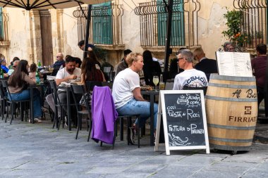 Noto, Sizilien - İtalya - 04-06-2025: İnsanlar Noto 'daki bir restoranın önünde masaya oturmuş yemek ve içeceklerin keyfini çıkarırken, girişte bir menü tahtası ve şarap fıçısı sergileniyor..
