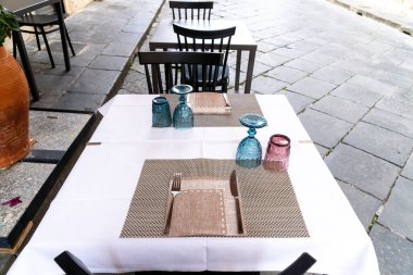 Noto, Sizilien - İtalya - 04-06-2025: Noto 'da iki kişilik açık restoran masası, renkli bardaklar ve çatal bıçaklar desenli örtülerin üzerine beyaz bir masa örtüsü yerleştirilmiş..
