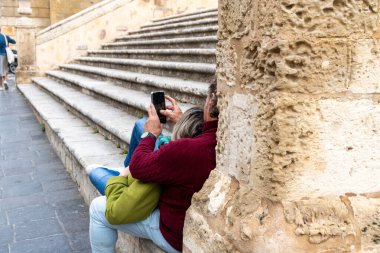 Noto, Sizilien - İtalya - 04-06-2025: Kırmızı kazaklı bir adam ve yeşil ceketli bir kadın taş bir duvara yaslanmış, birlikte akıllı telefona bakarken eski merdivenlerin yanında oturuyorlar..