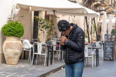 Noto, Sizilien - İtalya - 04-06-2025: Siyah ceketli ve şapkalı bir adam sessiz bir sokakta, boş sandalyeli ve kapalı şemsiyeli bir açık hava kafesinin önünde sigara yakıyor.