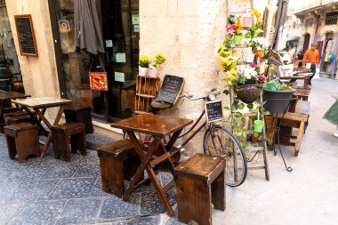 Syrakus, Sizilien - İtalya - 04-08-2025: Ahşap masalı kırsal sokak kafesi, çiçeklerle süslenmiş antika bir bisiklet ve Syracuse 'un tarihi merkezinde büyüleyici dar bir sokak manzarası.