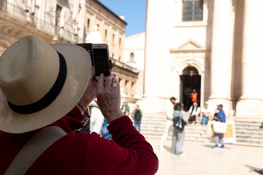 Syrakus, Sizilien - İtalya - 04-08-2025: Saman şapkalı turist akıllı bir telefonla Syracuse Katedrali 'ni fotoğraflıyor, güneşli taş basamaklarda toplanan insanları yakalıyor.