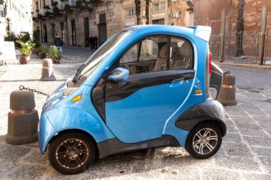 Syrakus, Sizilien - İtalya - 04-08-2025: Syrakus, Sicilya 'daki Syracus Katedrali' nin yakınına park edilmiş küçük mavi elektrikli araba, tarihi taş binalar ve saksı bitkileriyle çevrili..