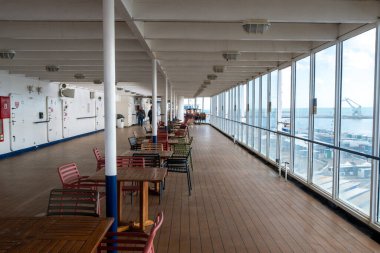 Cagliari - Sardinien - Italien - 03-15-2025: Ahşap döşeme, renkli sandalyeler, masalar ve liman manzarasına bakan geniş pencereli Europa Sarayı feribotunun üstü örtülü güvertesi.