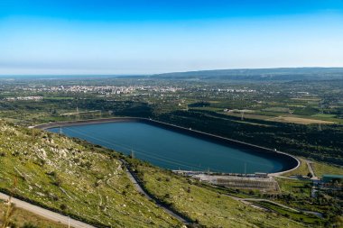 Pantalica, Sizilien - İtalya - 04-11-2025: Centrale Anapo yakınlarında bir tepenin altında, elektrik hatları, yollar, tarım arazileri ve mavi gökyüzü altındaki uzak şehir manzaralarıyla çevrili geniş bir rezervuar bulunuyor.